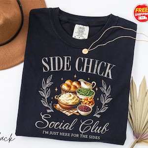 May include: Black crew neck sweatshirt with the text "SIDE CHICK Social Club" and the phrase "I'M JUST HERE FOR THE SIDES". The graphic features illustrations of Thanksgiving side dishes. A gold necklace is draped across the shirt.