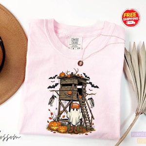 May include: A light pink Comfort Colors t-shirt featuring a Halloween-themed graphic. The design includes a wooden hunting blind, pumpkins, bats, and a ghost wearing a hunting hat. A necklace is draped across the shirt.