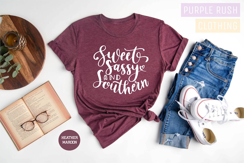 K&ouml;nnte beinhalten: Ein maronfarbenes T-Shirt aus Heather-Material mit dem Text "Sweet Sassy and Southern" in wei&szlig;en Buchstaben. Das Shirt liegt flach auf einer wei&szlig;en Oberfl&auml;che mit einem Buch, einer Brille und einer Jeans.