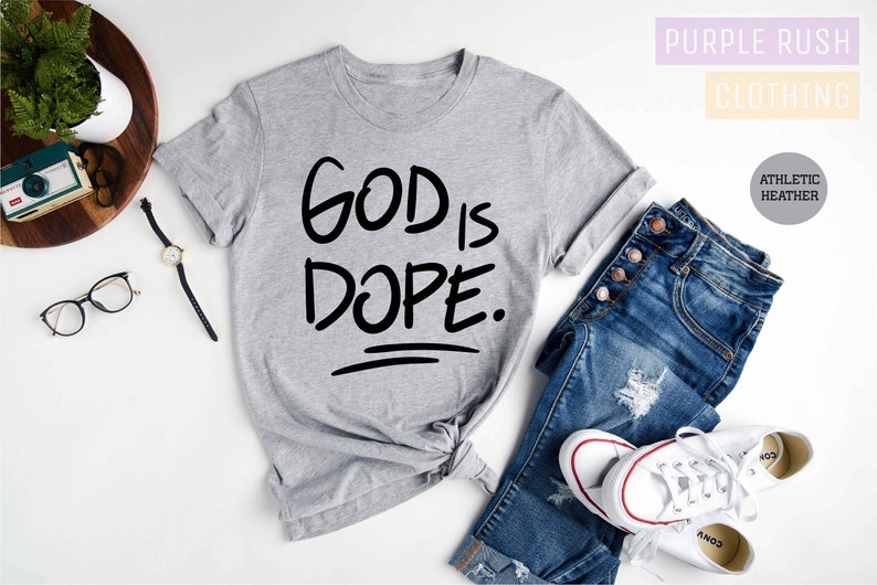 May include: Gray t-shirt with the text "GOD IS DOPE." printed in black. The shirt is shown on a white background with a pair of jeans and white sneakers.