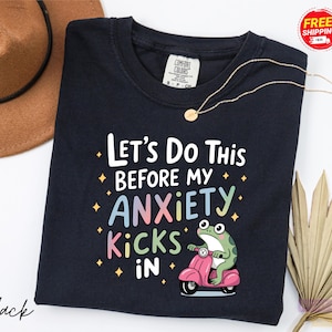 A black t-shirt with a colorful graphic design and text, along with a brown hat and a fan, all placed on a white background.