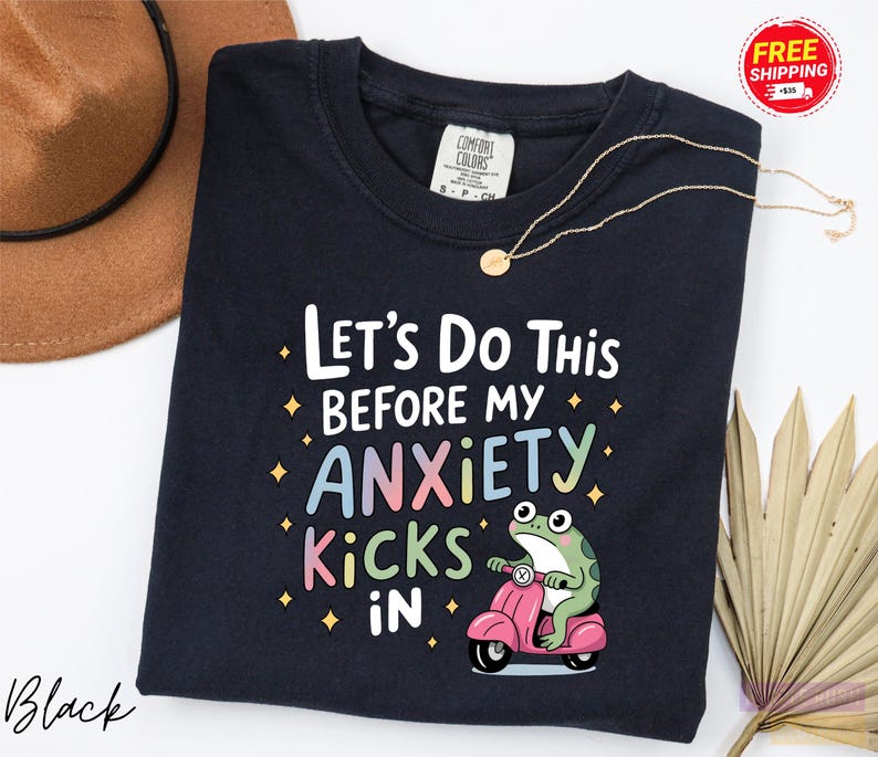 A black t-shirt with a colorful graphic design and text, along with a brown hat and a fan, all placed on a white background.