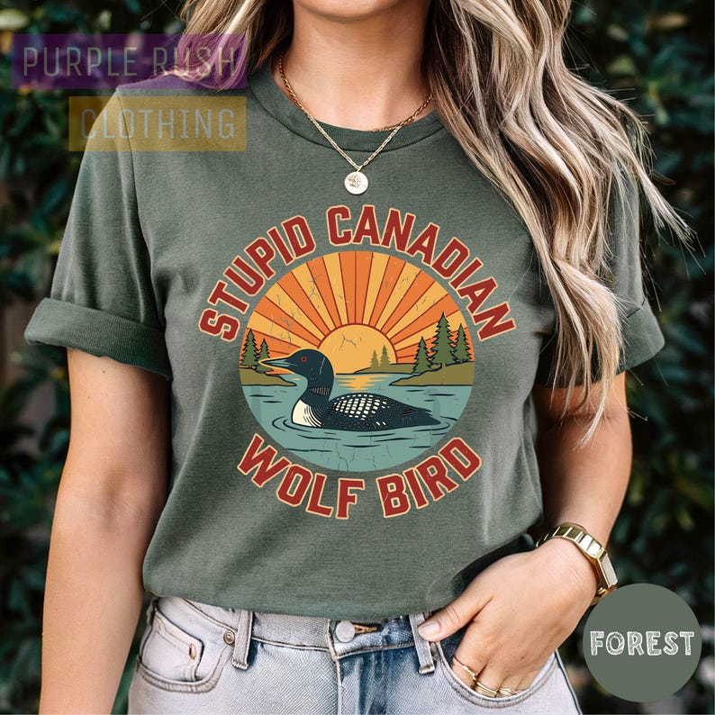 A woman wearing a green t-shirt with a graphic design of a duck and sun on it, along with the text "Stupid Canadian Wolf Bird" printed on it.