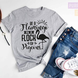 May include: A gray t-shirt with the text "Be a Flamingo in a Flock of Pigeons" printed in black. The shirt is shown on a white background with a pair of jeans and white sneakers.