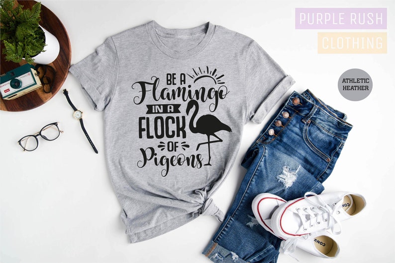 May include: A gray t-shirt with the text "Be a Flamingo in a Flock of Pigeons" printed in black. The shirt is shown on a white background with a pair of jeans and white sneakers.