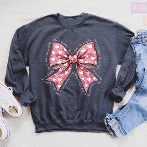 May include: Dark heather gray sweatshirt with a large pink bow design filled with white hearts. The words "You are beautiful, You are worthy, You are loved" are printed around the bow. White sneakers and blue jeans are also in the image.