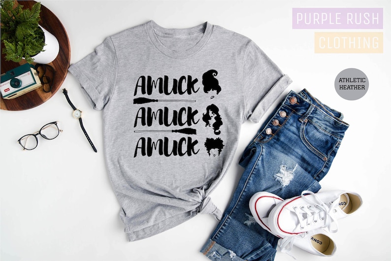May include: Athletic heather gray t-shirt with the text "AMUCK" and three witch silhouettes. The shirt is styled with blue jeans and white sneakers. The words "Purple Rush Clothing" are in the upper right corner.
