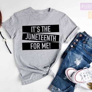 May include: A gray t-shirt with the text "It's the Juneteenth for me!" printed in black. The shirt is laid out on a white surface with a pair of jeans and white sneakers.
