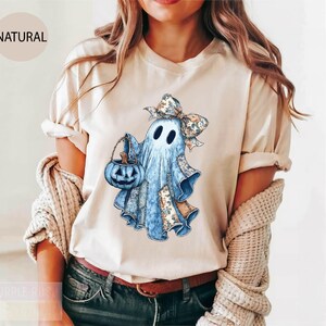 A woman wearing a white t-shirt with a graphic of a ghost holding a pumpkin and a bow.