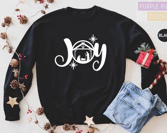 May include: Black sweatshirt with the word "Joy" in white script, featuring a nativity scene graphic. The sweatshirt is decorated with a Christmas theme, including pine cones, stars, and a red gift box. A pair of blue jeans is placed below the sweatshirt.