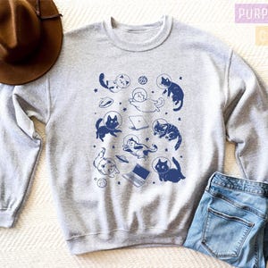 May include: A gray sweatshirt with a print of cats in space wearing astronaut helmets. The cats are in various poses, some floating, some holding laptops, and some holding a cup of coffee. The print is in shades of blue.