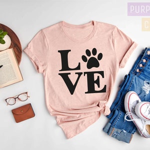 Pu&ograve; includere: Una maglietta rosa con la parola "LOVE" in lettere nere e un'impronta di zampa al centro della "O". La maglietta &egrave; stesa su una superficie bianca con un paio di jeans, scarpe da ginnastica bianche e un libro.
