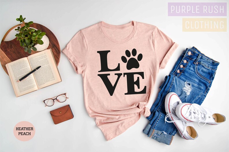 Pu&ograve; includere: Una maglietta rosa con la parola "LOVE" in lettere nere e un'impronta di zampa al centro della "O". La maglietta &egrave; stesa su una superficie bianca con un paio di jeans, scarpe da ginnastica bianche e un libro.