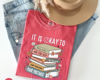 Puede incluir: Una camiseta rosa con el texto "It is okay to" y una pila de libros con frases como "Pedir ayuda", "Empezar de nuevo" y "Cometer errores".