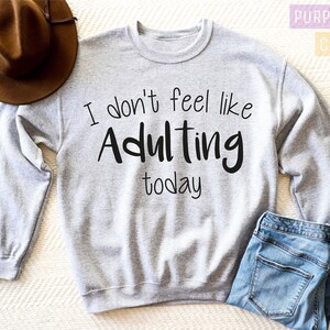 K&ouml;nnte beinhalten: Ein graues Sweatshirt mit dem Text "I don't feel like Adulting today" in Schwarz gedruckt.