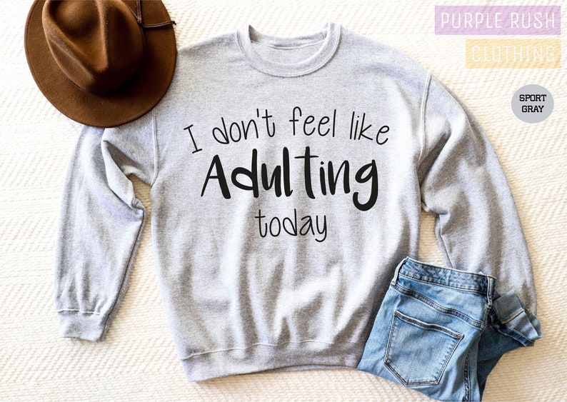 K&ouml;nnte beinhalten: Ein graues Sweatshirt mit dem Text "I don't feel like Adulting today" in Schwarz gedruckt.