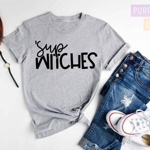 Puede incluir: Una camiseta gris jaspeada con la frase "'Sup Witches" en letras negras. La camiseta se combina con vaqueros azules y zapatillas blancas. La imagen tambi&eacute;n incluye un reloj, gafas y una peque&ntilde;a c&aacute;mara.