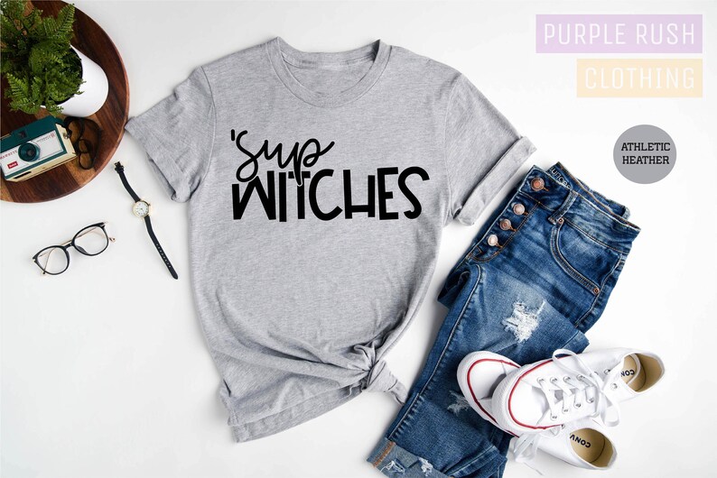 Puede incluir: Una camiseta gris jaspeada con la frase "'Sup Witches" en letras negras. La camiseta se combina con vaqueros azules y zapatillas blancas. La imagen tambi&eacute;n incluye un reloj, gafas y una peque&ntilde;a c&aacute;mara.