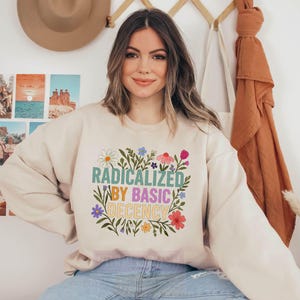 A woman wearing a white sweatshirt with a colorful graphic design and text, sitting in front of a wall with various photographs and a hat.