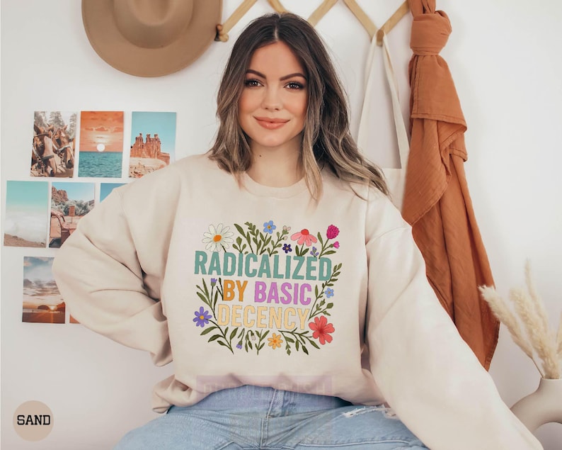 A woman wearing a white sweatshirt with a colorful graphic design and text, sitting in front of a wall with various photographs and a hat.