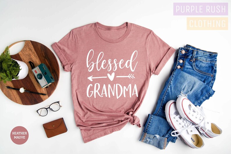 May include: A heather mauve t-shirt with the text "blessed grandma" in white lettering with an arrow and heart graphic. The shirt is laid flat on a white background with a pair of glasses, a wooden chopping board, a plant, a camera, a brown wallet, and a pair of blue jeans with white trainers.