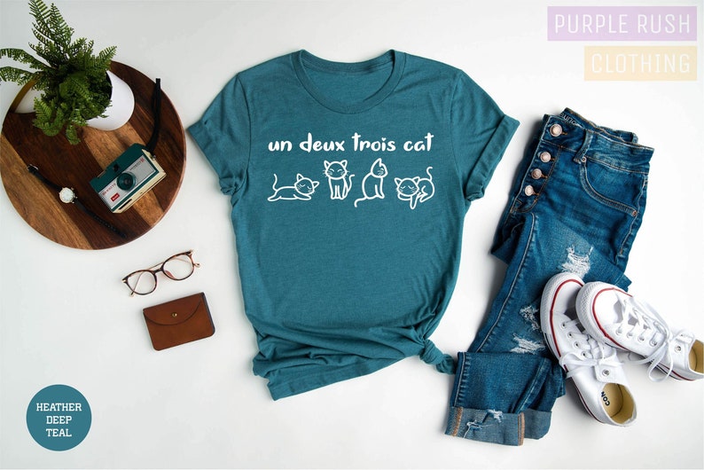 May include: Teal blue t-shirt with white text that says "un deux trois cat" and a row of four white cartoon cats.