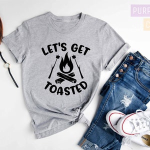 May include: A heather gray t-shirt with the text "LET'S GET TOASTED" in black, featuring a campfire graphic. The shirt is paired with blue jeans and white sneakers. The words "Purple Rush Clothing" are in the background.