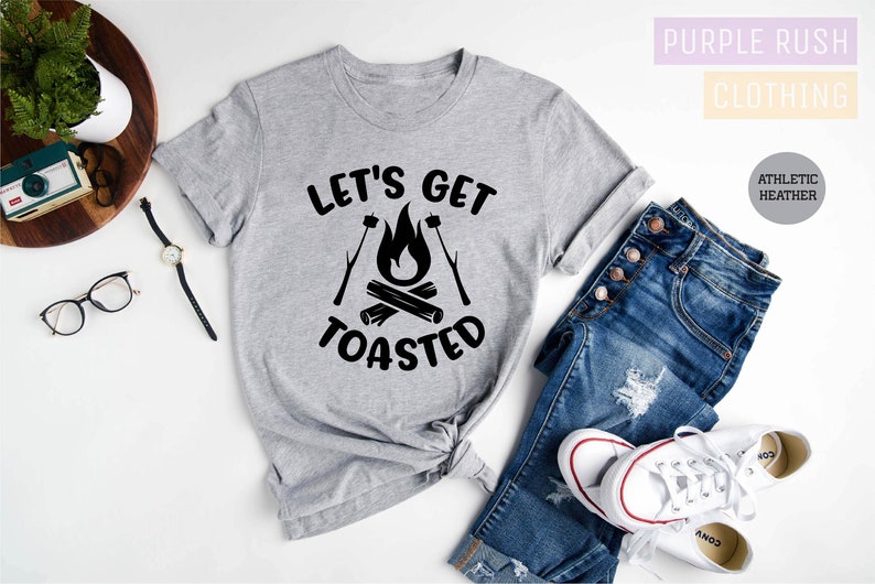 May include: A heather gray t-shirt with the text "LET'S GET TOASTED" in black, featuring a campfire graphic. The shirt is paired with blue jeans and white sneakers. The words "Purple Rush Clothing" are in the background.