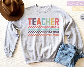 Op de afbeelding: Een grijs sweatshirt met het woord "TEACHER" in regenboogkleuren en de woorden "BELIEVER", "EDUCATOR", "INNOVATOR" en "MOTIVATOR" eronder. Het sweatshirt heeft een geruit patroon achter de tekst.