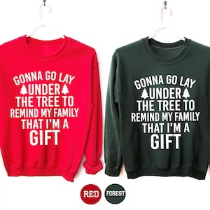 May include: Two sweatshirts, one red and one forest green, each with the text "GONNA GO LAY UNDER THE TREE TO REMIND MY FAMILY THAT I'M A GIFT" in white. The sweatshirts are hanging on white hangers.