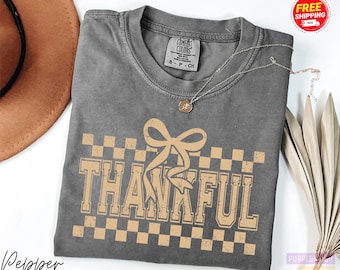 May include: A gray t-shirt with the word "THANKFUL" in a tan, vintage-style font, with a bow graphic above the word. The shirt has a checkered pattern and a round neckline. A silver necklace with a pendant is on the shirt.