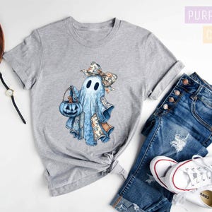 a casual outfit consisting of a gray t-shirt with a ghost design, a pair of blue jeans, and white sneakers. The outfit is laid out on a white background, with a pair of glasses and a camera visible as well.