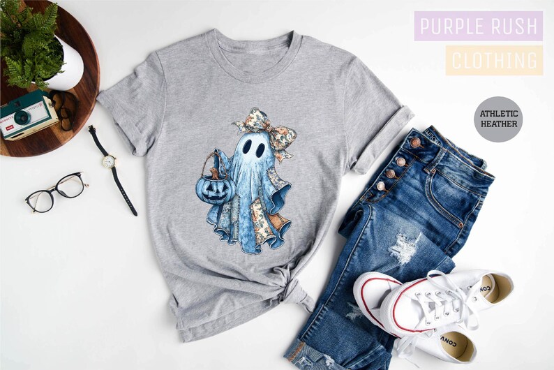 a casual outfit consisting of a gray t-shirt with a ghost design, a pair of blue jeans, and white sneakers. The outfit is laid out on a white background, with a pair of glasses and a camera visible as well.