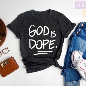 May include: Dark gray heather t-shirt with the text "GOD IS DOPE." printed in white. The shirt is laid flat on a white surface with a pair of jeans, a wallet, a camera, and a potted plant.
