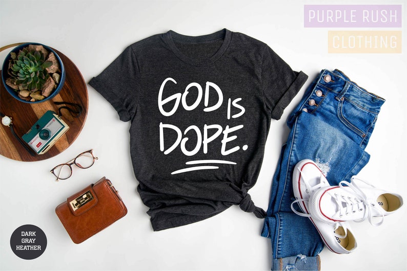 May include: Dark gray heather t-shirt with the text "GOD IS DOPE." printed in white. The shirt is laid flat on a white surface with a pair of jeans, a wallet, a camera, and a potted plant.