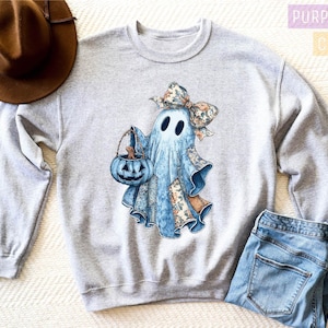 a gray sweatshirt with a graphic of a ghost wearing a bow and holding a pumpkin, along with a brown hat and a pair of blue jeans.