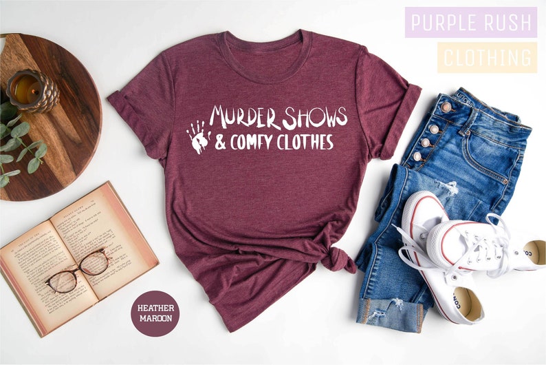 May include: A maroon t-shirt with the text "Murder Shows & Comfy Clothes" in white. The shirt is shown on a white background with a book, glasses, and a pair of jeans.