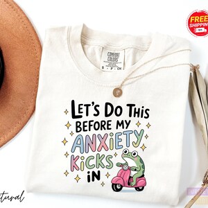 A white t-shirt with a colorful graphic design featuring a frog riding a pink scooter and the text "Let's Do This Before My Anxiety Kicks In".
