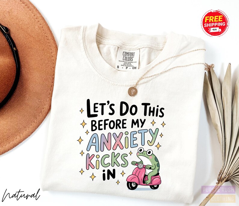 A white t-shirt with a colorful graphic design featuring a frog riding a pink scooter and the text "Let's Do This Before My Anxiety Kicks In".