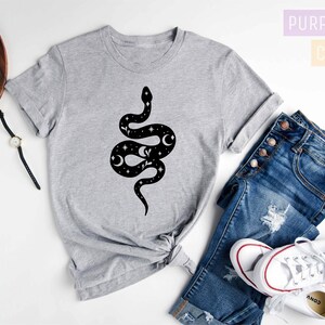 May include: A gray t-shirt with a black snake design featuring stars and moons. The shirt is on a white surface with a pair of jeans, a watch, and a camera.