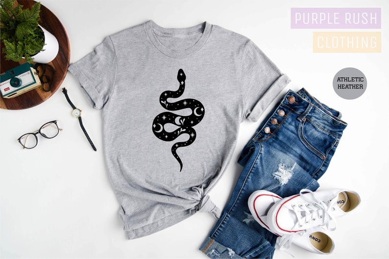 May include: A gray t-shirt with a black snake design featuring stars and moons. The shirt is on a white surface with a pair of jeans, a watch, and a camera.
