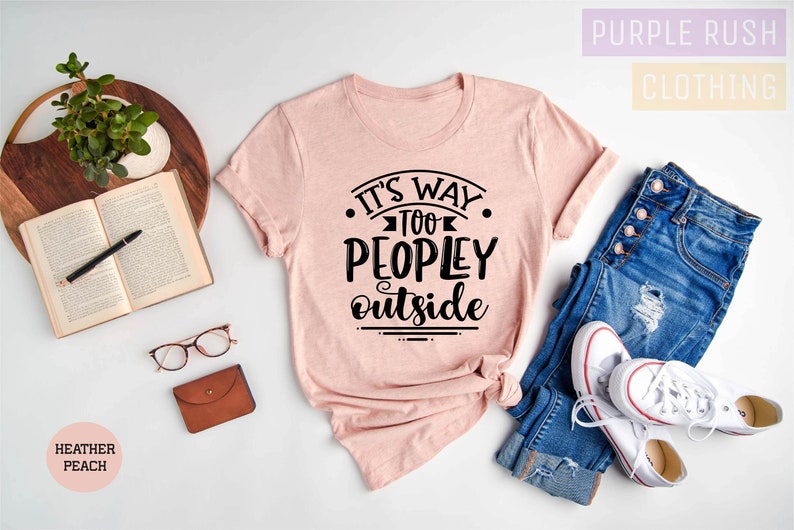 May include: A pink t-shirt with the text "It's Way Too Peopley Outside" in black lettering. The shirt is laid out on a white surface with a pair of jeans and white sneakers.