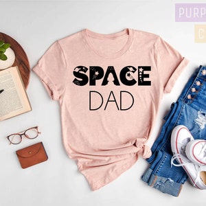 K&ouml;nnte beinhalten: Ein pinkfarbenes T-Shirt mit dem Text "SPACE DAD" in schwarzen Buchstaben. Die Buchstaben sind mit Sternen und Planeten verziert. Das Shirt liegt auf einer wei&szlig;en Oberfl&auml;che mit einer Jeans und wei&szlig;en Turnschuhen.