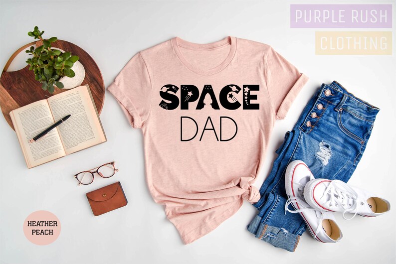 K&ouml;nnte beinhalten: Ein pinkfarbenes T-Shirt mit dem Text "SPACE DAD" in schwarzen Buchstaben. Die Buchstaben sind mit Sternen und Planeten verziert. Das Shirt liegt auf einer wei&szlig;en Oberfl&auml;che mit einer Jeans und wei&szlig;en Turnschuhen.