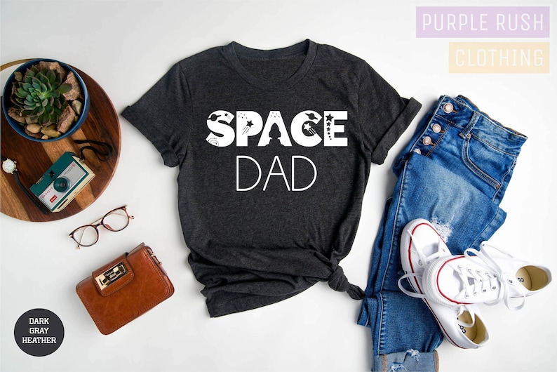 K&ouml;nnte beinhalten: Dunkelgraues T-Shirt mit wei&szlig;em Text "SPACE DAD" und einer Grafik mit einer Rakete, Sternen und Planeten.