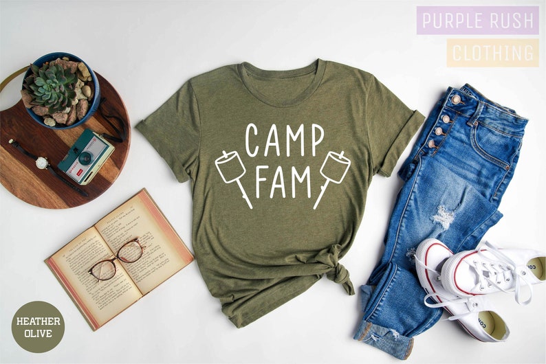 May include: A green t-shirt with the text "Camp Fam" and a graphic of two marshmallows on sticks. The shirt is laid flat on a white surface with a pair of jeans, a book, and a plant in a pot.