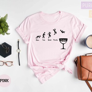 Op de afbeelding: Roze T-shirt met een afbeelding van een stokfiguur voor elke dag van de week, waarbij de figuur voor vrijdag een glas wijn vasthoudt. Op het glas staat de tekst "Friday!" Het shirt ligt plat op een wit oppervlak met een plant, een bril, een horloge, een handtas en een kopje koffie.