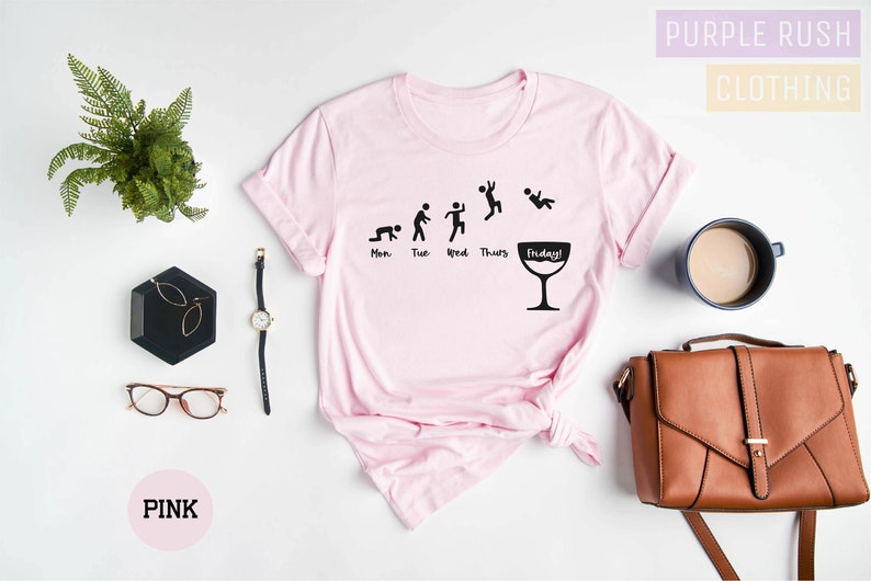 Op de afbeelding: Roze T-shirt met een afbeelding van een stokfiguur voor elke dag van de week, waarbij de figuur voor vrijdag een glas wijn vasthoudt. Op het glas staat de tekst "Friday!" Het shirt ligt plat op een wit oppervlak met een plant, een bril, een horloge, een handtas en een kopje koffie.