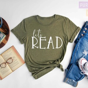 May include: A heather olive green t-shirt with the text "let's READ" in white lettering. The shirt is shown on a white background with a book, glasses, a potted plant, a camera, and a pair of blue jeans with white trainers.