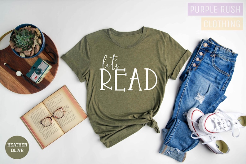 May include: A heather olive green t-shirt with the text "let's READ" in white lettering. The shirt is shown on a white background with a book, glasses, a potted plant, a camera, and a pair of blue jeans with white trainers.